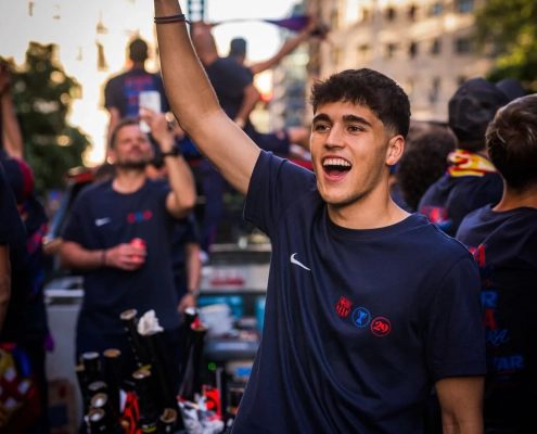 Person in a dark blue sports jersey with logos, celebrating outdoors with buildings and people in the background.
