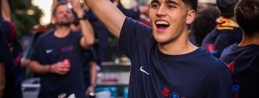 Person in a dark blue sports jersey with logos, celebrating outdoors with buildings and people in the background.